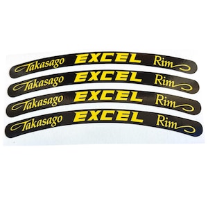 Puede incluir: Cuatro calcomanías negras curvas con el texto "Takasago EXCEL Rim" en amarillo. Las calcomanías están diseñadas para una llanta de motocicleta o bicicleta. El fondo es blanco.