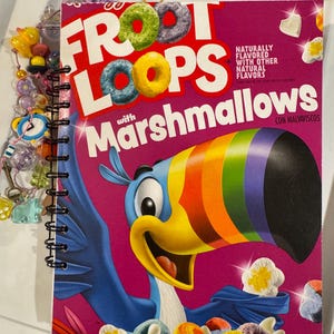 May include: A spiral-bound notebook featuring a vibrant cover design of a Froot Loops cereal box. The cover displays the cereal's logo, a toucan, and colorful cereal pieces. A beaded charm hangs from the spiral binding.