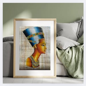 May include: Framed artwork depicting a profile of an Egyptian queen, featuring a blue and gold headdress and ornate collar. The artwork is set against a neutral background, framed in light wood, and placed near a bed with pillows and a green blanket.
