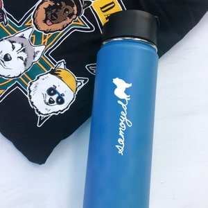 Könnte beinhalten: Eine blaue, isolierte Wasserflasche mit schwarzem Deckel und einer weißen Samojeden-Hundesilhouette und Text. Im Hintergrund ein schwarzes T-Shirt mit Hunde-Gesichtern.