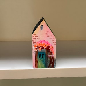 Puede incluir: Una pequeña casa de madera pintada de rosa con una puerta azul y un arco floral. La casa está decorada con una ventana amarilla y detalles de ladrillo.