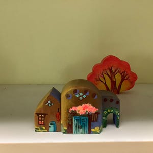 May include: Hand-painted wooden village set featuring three buildings and a tree. The buildings have arched roofs, painted doors, and windows. The tree is red and orange. The set is displayed on a white shelf against a yellow background.