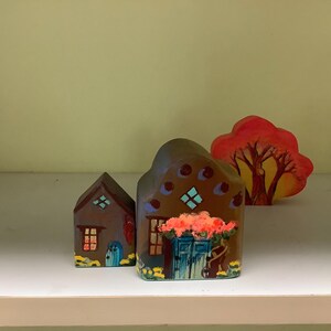 May include: Hand-painted wooden figurines of a house and a tree. The house figurines are brown with blue doors and windows, and the tree is red and orange. The house has a flower garden painted on the front.