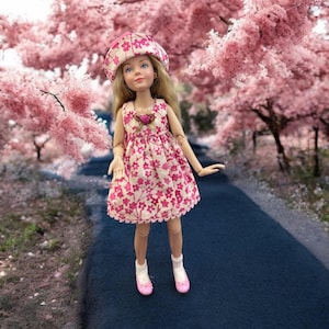 May include: A doll wearing a pink and white floral dress and a pink hat stands on a path in a cherry blossom tree grove.
