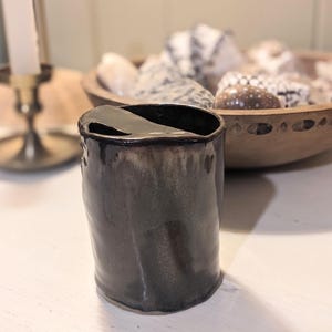 May include: A cylindrical ceramic cup with a dark, glossy, metallic-toned glaze. The rim features a distinct, asymmetrical cutout or notch on one side, creating an uneven edge. The surface has a smooth, reflective finish with subtle color variations.