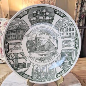 May include: A white commemorative plate with a green border and illustrations of buildings and text. The plate commemorates the Salem Evangelical Lutheran Church's bicentennial in 1975. The plate is displayed on a gold stand.