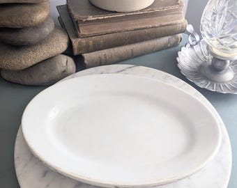 May include: A plain white oval ceramic platter resting on a circular white marble board. The platter features a smooth, glossy finish with a simple rim, providing a clean and neutral surface for serving food items in a dining or kitchen setting.