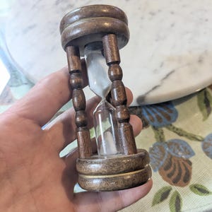 May include: A dark brown wooden hourglass featuring a clear glass bulb filled with white sand. The frame consists of two circular end pieces connected by four vertical, turned wooden pillars that support the central glass timer mechanism.