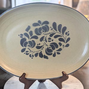 May include: An oval-shaped ceramic platter with a light beige, speckled finish and a dark blue rim. The center of the platter features a dark blue, hand-painted floral motif consisting of stylized leaves, petals, and small circular dots.