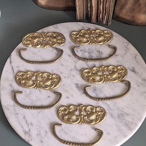 May include: Six antique brass drawer pulls and backplates arranged on a white marble surface. The backplates have an ornate, symmetrical design, and the pulls are curved with a textured pattern. The hardware is likely for furniture restoration.