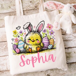 May include: A white canvas tote bag featuring a yellow chick wearing bunny ears, surrounded by colourful Easter eggs and flowers. The name "Sophia" is written in pink below the chick.