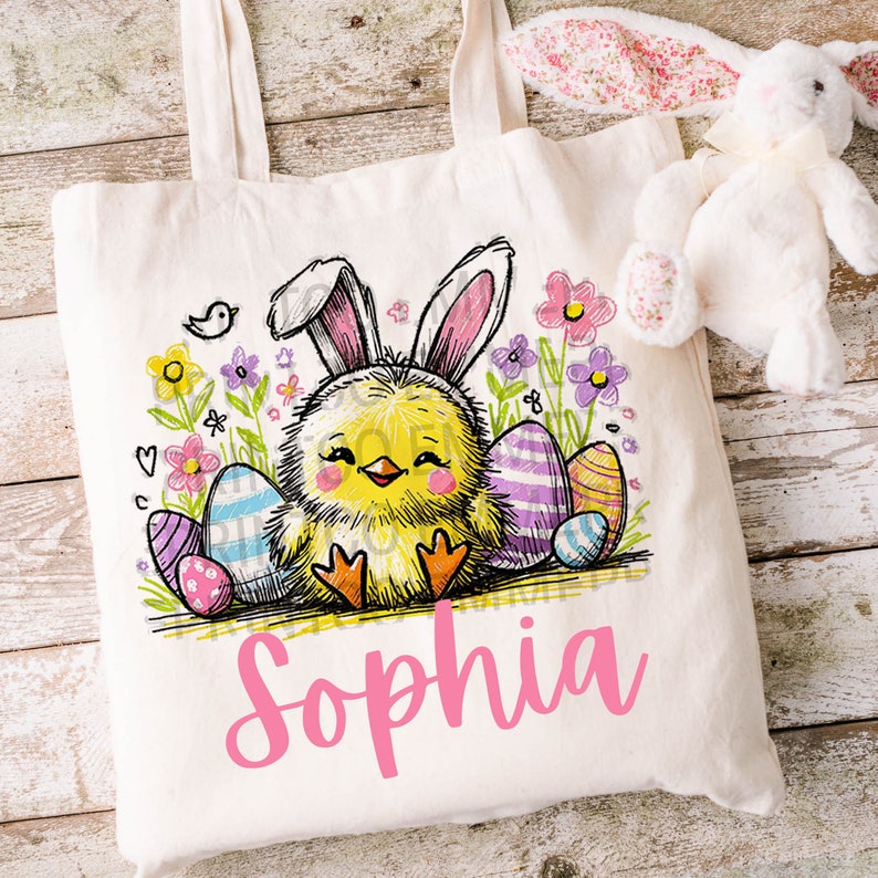 May include: A white canvas tote bag featuring a yellow chick wearing bunny ears, surrounded by colourful Easter eggs and flowers. The name "Sophia" is written in pink below the chick.