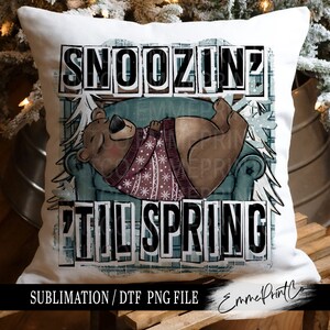 May include: A white pillow with a brown bear wearing a red and white sweater sleeping in a blue armchair. The pillow has the text "Snoozin' 'til Spring" in black letters.