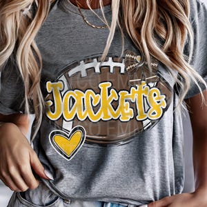 May include: Gray t-shirt with a brown and white football graphic. The football has the word "Jackets" written on it in yellow letters. There is a yellow heart on the bottom of the football.