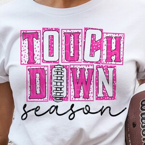 Puede incluir: Camiseta blanca con un gráfico rosa y blanco que dice "Touchdown Season" con un gráfico de fútbol en el medio.