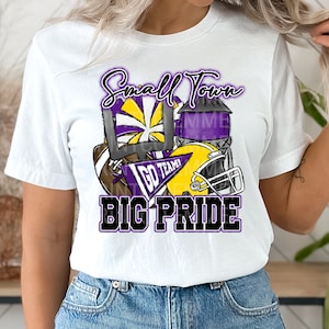 May include: White t-shirt with a purple, yellow, and black graphic design. The graphic features a football helmet, a football, a cheerleader's pom-pom, and a water tower. The text "Small Town Big Pride" is written in black with a purple outline.