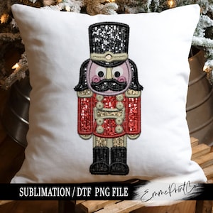 May include: A red and black nutcracker with a sparkly black hat and a gold trim. The nutcracker has a pink face with black eyes and a black moustache. The nutcracker is on a white cushion with the text "SUBLIMATION / DTF PNG FILE" and "EmmePrintCo" on the bottom.