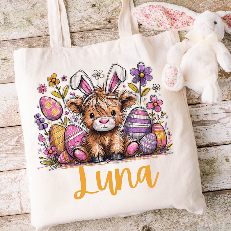 May include: A white canvas tote bag featuring a brown and white cartoon Highland cow wearing pink bunny ears. The cow is surrounded by colourful Easter eggs and flowers. The name "Luna" is written in yellow at the bottom of the bag.