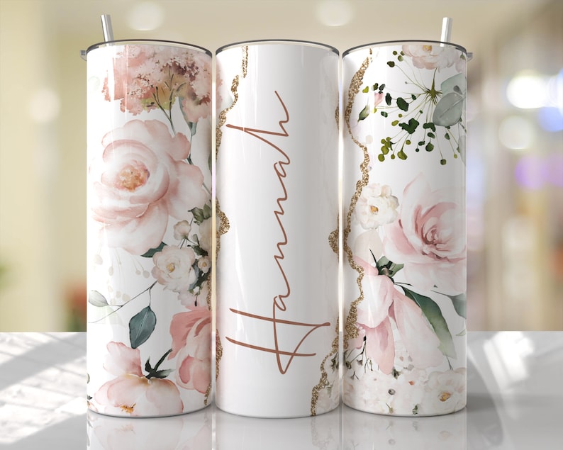 Puede incluir: Tres vasos de acero inoxidable con un dise&ntilde;o floral. El vaso del centro tiene el nombre "Hannah" escrito en letras doradas. Los vasos est&aacute;n sobre una superficie blanca.