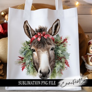 Puede incluir: Una ilustración de acuarela de un burro con un lazo rojo y una corona de Navidad. El burro está mirando directamente al espectador. La imagen está en una bolsa de mano blanca con el texto "SUBLIMATION PNG FILE" y "EmmePrintCo" en la parte inferior.