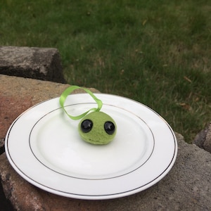 以下が含まれることがあります： 金色の縁取りのある白い皿の上に、黒い目をした小さな緑色のフェルトの生き物が乗っています。生き物は緑色のリボンに繋がっています。