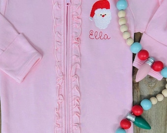 May include: Pink baby footed pajamas with a Santa Claus face embroidered on the chest, with the name "Ella" in red script. The pajamas have a ruffled trim down the front and a full zipper. A wooden Christmas light garland is draped nearby.