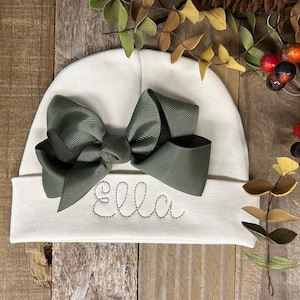 May include: A white knitted baby hat with a large olive green bow and the name "Ella" embroidered on the front.