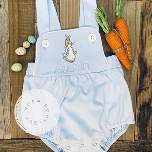 May include: Light blue baby romper with white trim, featuring an embroidered Peter Rabbit design and the name "Austin". Includes decorative carrots and speckled Easter eggs. The romper has button closures and adjustable straps.