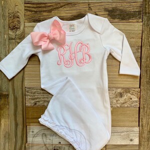 May include: White baby gown with long sleeves, a pink bow, and a pink embroidered monogram. The gown has a ruffled hem and the text "RB Blanks" and "Nora Boutique" are visible.