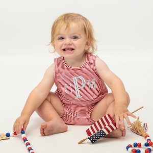 Op de afbeelding: Een baby draagt een rood-wit gestreept rompertje met de letters "CPM" geborduurd op de voorkant. De baby zit op een wit oppervlak en speelt met een rood-wit-blauwe kralenketting en een kleine Amerikaanse vlag.