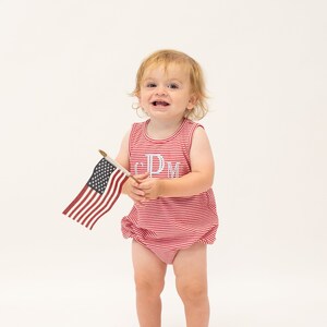 Op de afbeelding: Een jong kind draagt een rood-wit gestreept rompertje met de letters "CPM" geborduurd op de voorkant, houdt een kleine Amerikaanse vlag vast.