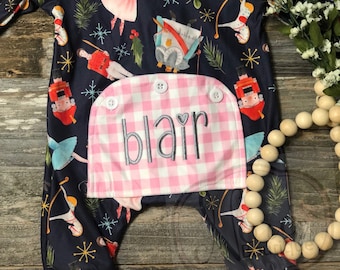 May include: Navy blue baby romper with a pink and white gingham pocket embroidered with the name "Blair". The romper features a Nutcracker-themed print with ballerinas, soldiers, and snowflakes. It has a ruffled collar and cuffs.