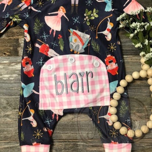May include: Navy blue baby romper with a pink and white gingham pocket embroidered with the name "Blair". The romper features a Nutcracker-themed print with ballerinas, soldiers, and snowflakes. It has a ruffled collar and cuffs.