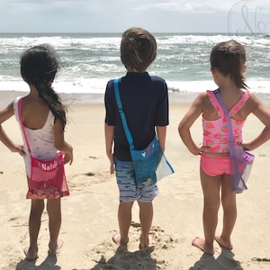 May include: Three children on a beach, wearing colorful swimsuits and carrying mesh bags with their names on them. The bags are pink, blue, and purple.
