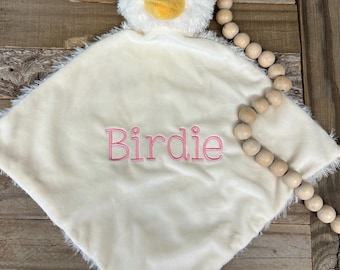May include: A soft, cream-colored plush security blanket featuring a stuffed duck head with a yellow beak and black eyes. The center of the square blanket is embroidered with the name Birdie in pink thread, and the edges have a fuzzy texture.