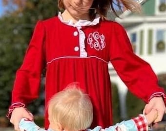 May include: Two children in matching outfits stand on a grassy lawn. One child wears a red dress with white trim and a monogram. The other child wears blue Santa-themed pajamas with the name "BAKER" on the back. A "NORA BOUTIQUE" logo is visible.