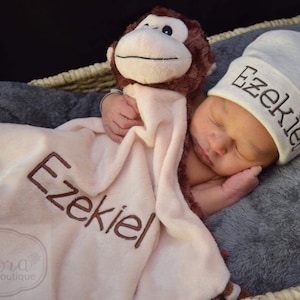 May include: A light pink security blanket with a brown plush monkey head and the name Ezekiel embroidered in dark brown thread, paired with a matching light gray beanie hat featuring the name Ezekiel embroidered in dark brown thread.