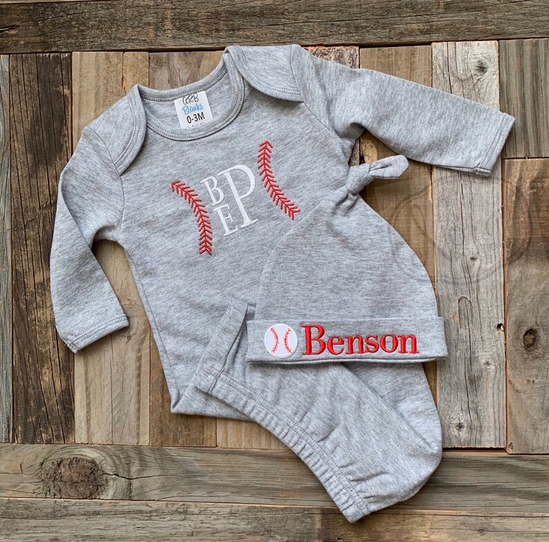 May include: Gray baby onesie and hat set with red baseball stitching and the letters "B.E.P." on the onesie. The hat has a red baseball graphic and the name "Benson" embroidered on it.