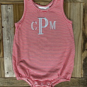 Op de afbeelding: Rode en witte gestreepte baby romper met de letters "CPM" geborduurd op de voorkant. De romper heeft een gerimpelde zoom en is gemaakt van een zachte, comfortabele stof.
