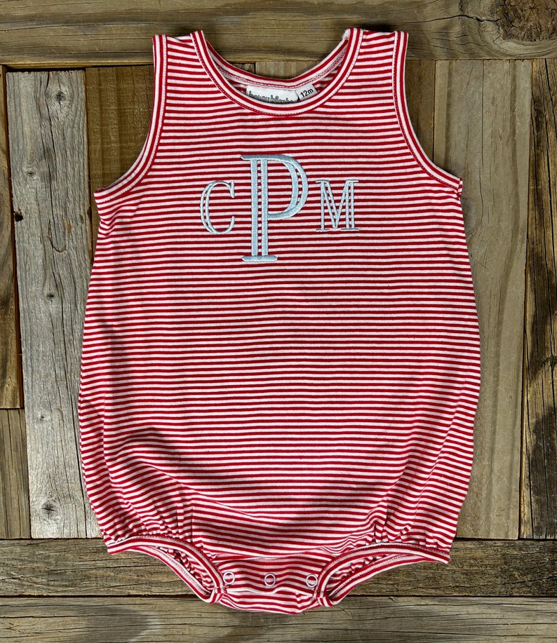 Op de afbeelding: Rode en witte gestreepte baby romper met de letters "CPM" geborduurd op de voorkant. De romper heeft een gerimpelde zoom en is gemaakt van een zachte, comfortabele stof.