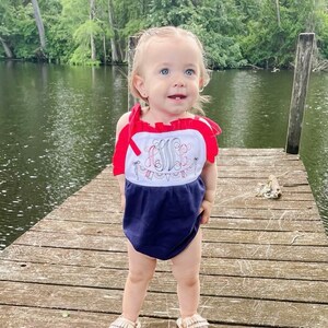 Puede incluir: Una ni&ntilde;a peque&ntilde;a con un mameluco azul y blanco sin mangas con ribetes rojos y un monograma bordado blanco. El mameluco tiene un dise&ntilde;o de banderines rojo, blanco y azul. La ni&ntilde;a est&aacute; de pie en un muelle de madera.
