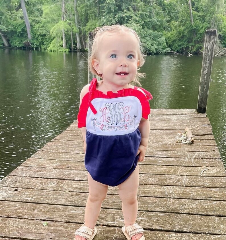 Puede incluir: Una ni&ntilde;a peque&ntilde;a con un mameluco azul y blanco sin mangas con ribetes rojos y un monograma bordado blanco. El mameluco tiene un dise&ntilde;o de banderines rojo, blanco y azul. La ni&ntilde;a est&aacute; de pie en un muelle de madera.