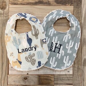 May include: Two baby bibs with a cactus and llama design. One bib is white with a blue and brown cactus and llama design and the name "Landry" embroidered on it. The other bib is gray with a white cactus design and the initials "LRH" embroidered on it.