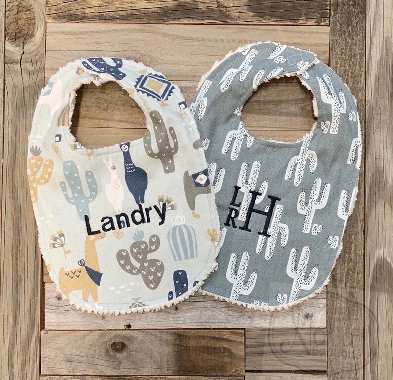 May include: Two baby bibs with a cactus and llama design. One bib is white with a blue and brown cactus and llama design and the name "Landry" embroidered on it. The other bib is gray with a white cactus design and the initials "LRH" embroidered on it.