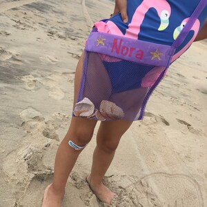 May include: A purple mesh bag with a pink flamingo design and the name "Nora" embroidered on the front. The bag is worn by a child on a sandy beach.