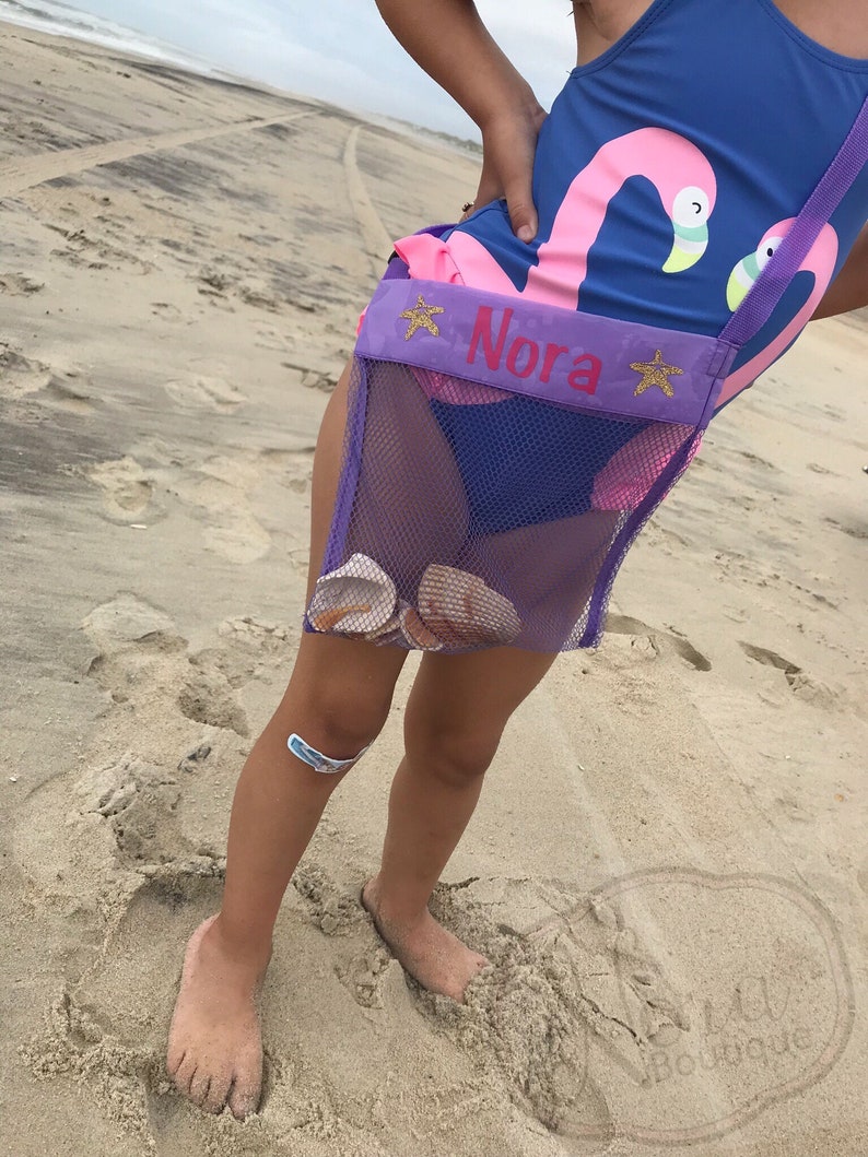 May include: A purple mesh bag with a pink flamingo design and the name "Nora" embroidered on the front. The bag is worn by a child on a sandy beach.