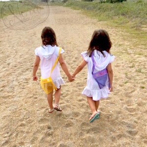 May include: Two young girls wearing white dresses with colorful mesh bags walk along a sandy path. The girl in the front is wearing a yellow bag with a flower design. The girl in the back is wearing a purple bag with the name "Nora" embroidered on it.