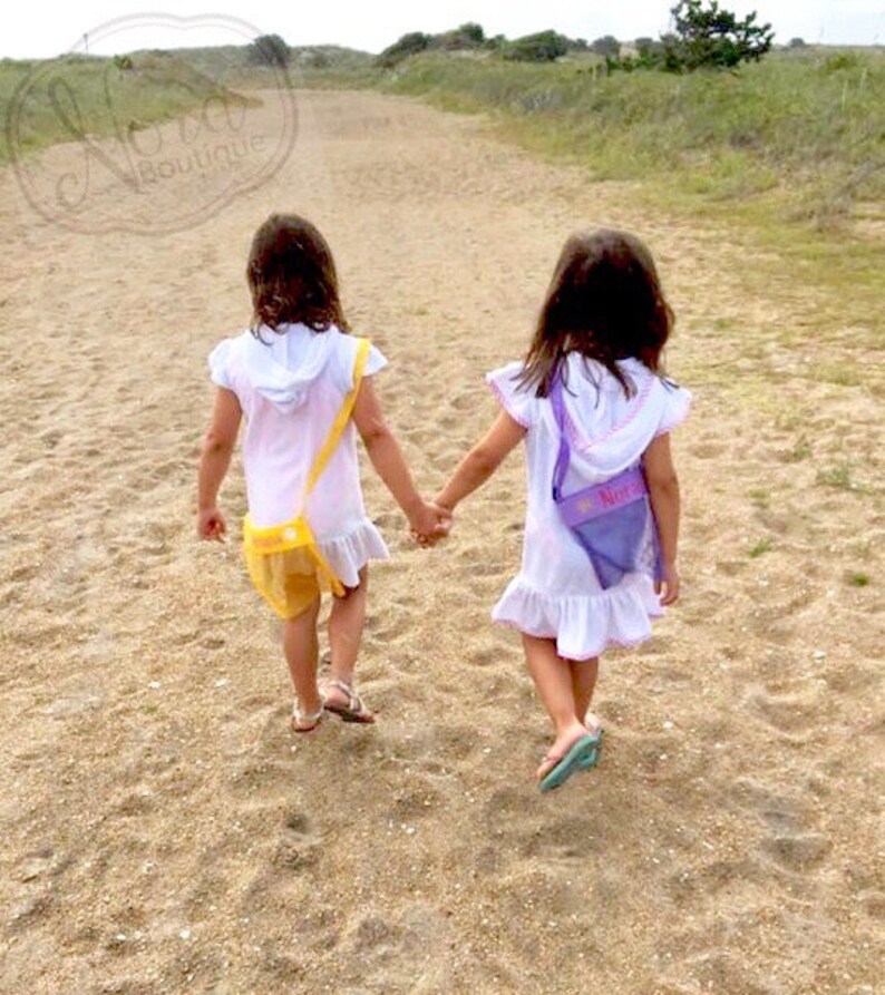 May include: Two young girls wearing white dresses with colorful mesh bags walk along a sandy path. The girl in the front is wearing a yellow bag with a flower design. The girl in the back is wearing a purple bag with the name "Nora" embroidered on it.