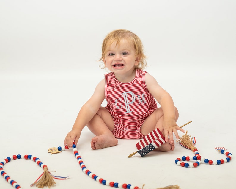 Op de afbeelding: Een baby draagt een rood-wit gestreept rompertje met de letters "CPM" geborduurd op de voorkant. De baby zit op een wit oppervlak en speelt met een rood-wit-blauwe kralenketting en een kleine Amerikaanse vlag.