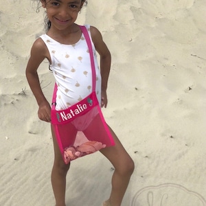 May include: A young girl wearing a white swimsuit with gold palm tree print is holding a pink mesh beach bag with the name "Natalie" embroidered on it. The bag is filled with seashells.